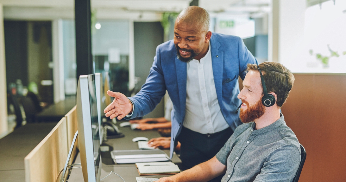 Call center manager explaining something to an agent at their desk. They are both smiling and looking at a computer screen.