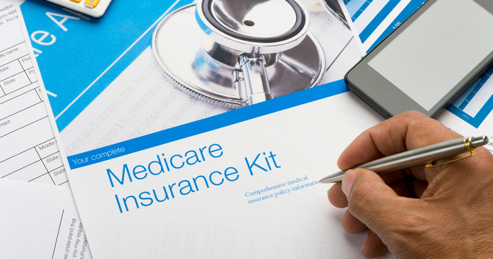 A hand holding a pen above a medicare health insurance kit, with a cell phone resting on the table beside it.