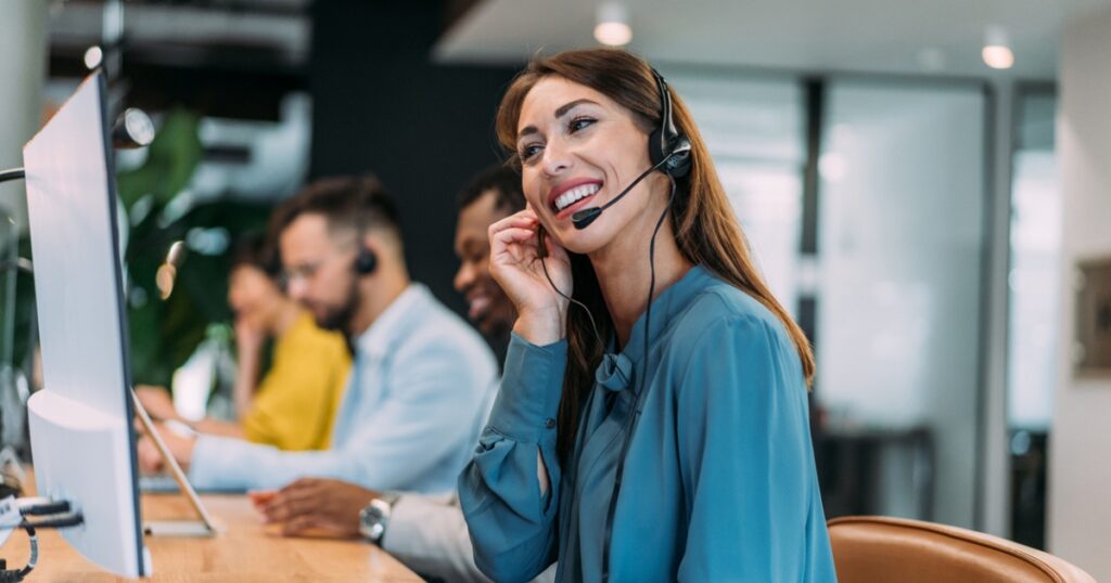 A call center agent wearing a headset is smiling while working at a computer in a modern office. Two other colleagues are working in the background, also using headsets.