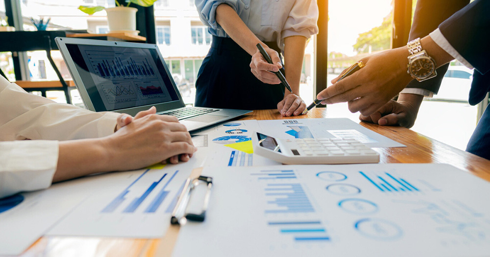 A group of call center professionals collaborating at a desk with charts, graphics, and a laptop displaying statistics and trends. They are discussing business strategies.