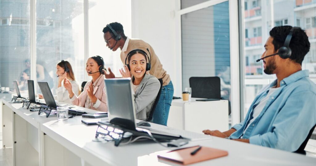 Group of call center agents wearing headsets and making calls at the same time. A few agents are smiling and talking to each other.