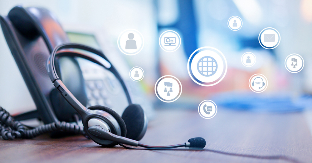 Headset next to a landline phone on a desk. Above the desk there are floating icons that represent Voice over Internet Protocol (VoIP) technology.