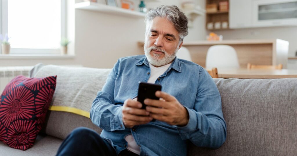 A middle-aged man sitting on a couch in a bright living room, looking at his cellphone.