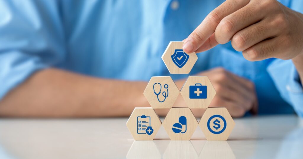 A close-up of a hand placing wooden blocks with healthcare-related icons, including a shield, stethoscope, and pill, into a pyramid structure.