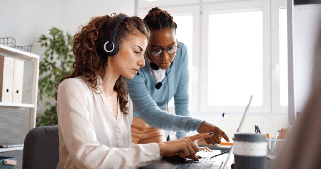 Two call center employees are collaborating at a desk, both wearing headsets. One is leaning over and pointing at a laptop screen, explaining something to the other.