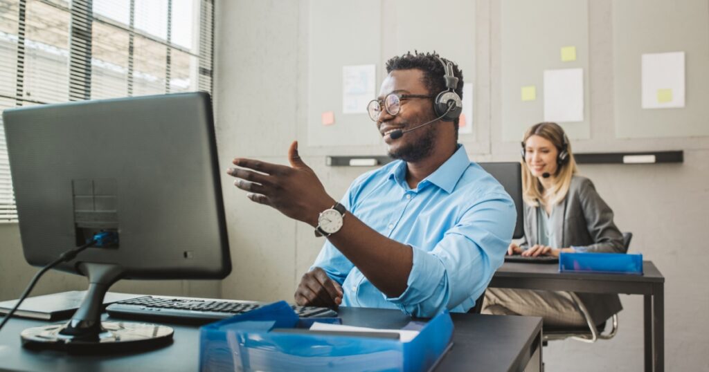 A sales agent wearing a headset and smiling while on a call. A colleague is working in the background.