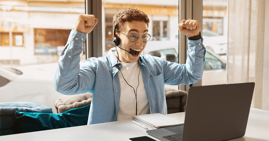 A young man wearing a headset raises his fists in excitement while looking at a laptop, symbolizing a successful outbound sales call.