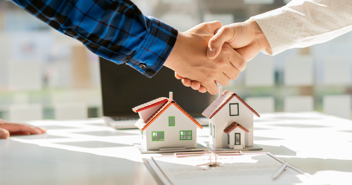 Two people shake hands over a table with miniature houses after closing a real estate wholesaling deal.