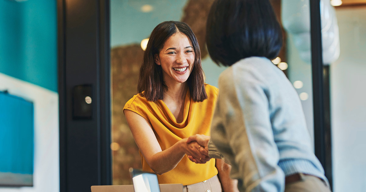 Two women smiling and shaking hands in an office setting, representing the relationship-building and verification steps involved in the Know Your Customer (KYC) process in telecom.