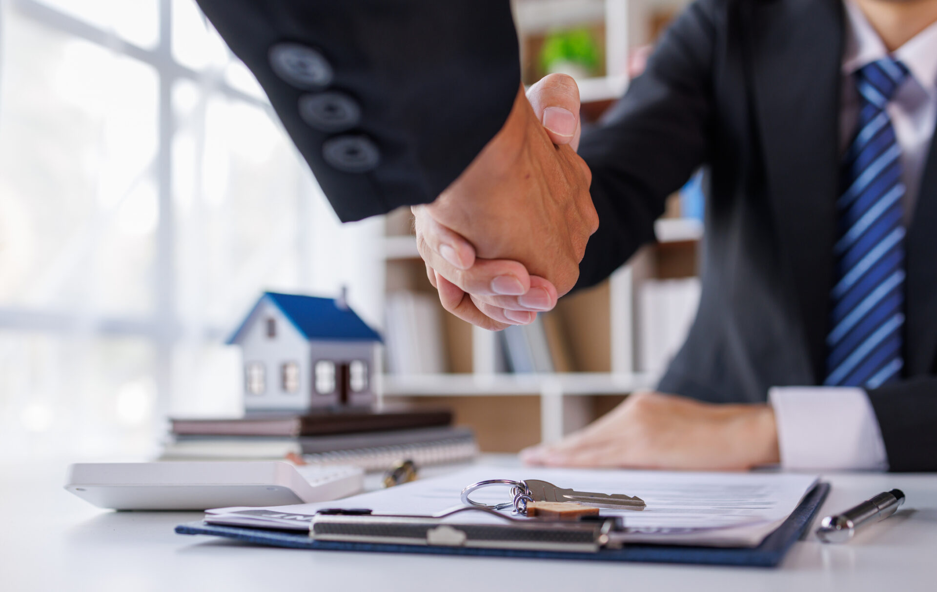 Two business professionals in suits shaking hands over a real estate contract, with a model house, keys, and documents on the desk.