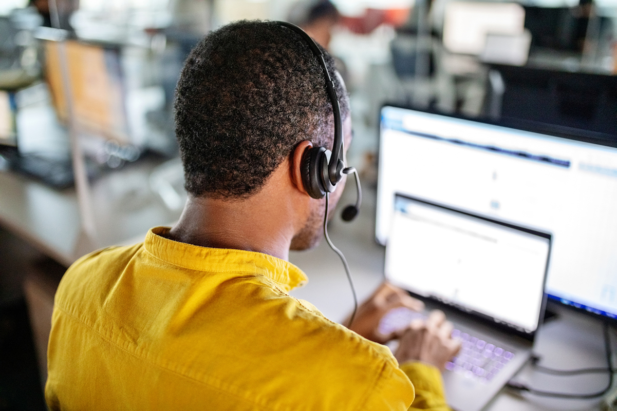 Salesman wearing a headset and sitting at his desk. He is making phone calls using cold calling software on both his laptop and monitor.
