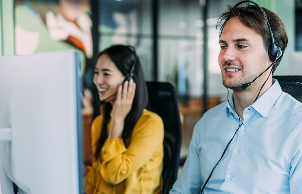 Two smiling insurance agents wearing headsets and sitting at their desks while making phone calls to potential clients.