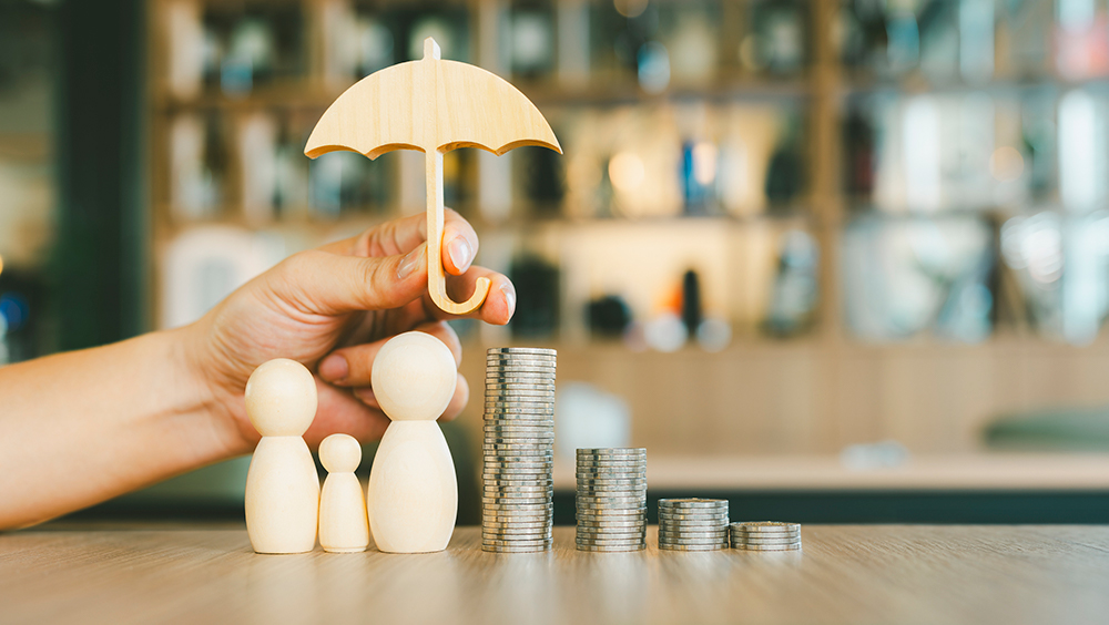Hand holding a wooden umbrella over wooden figures representing a family beside stacks of coins, symbolizing financial protection and insurance.